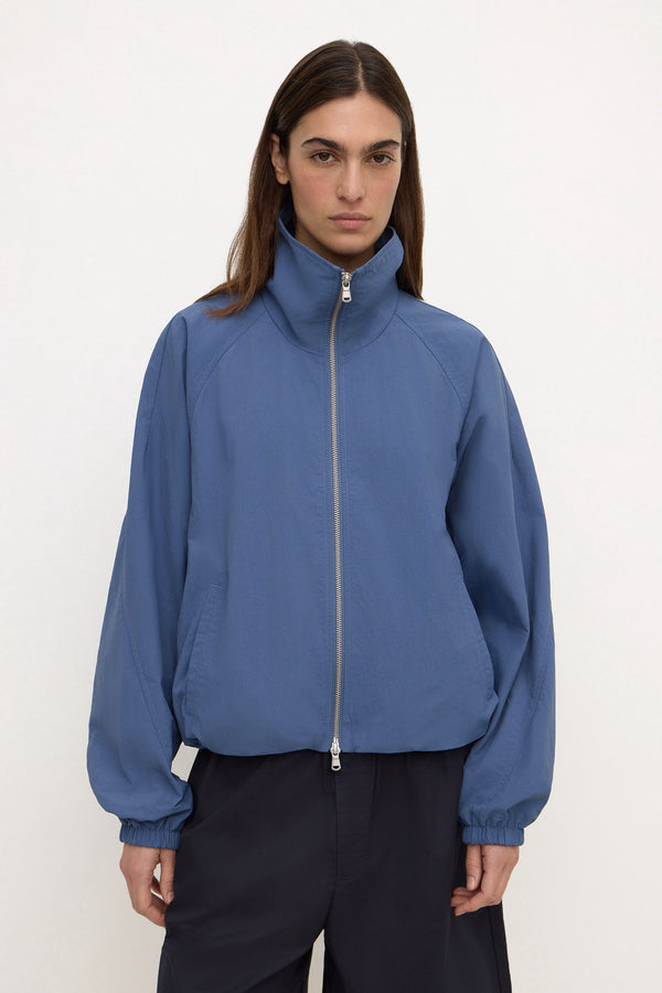 A woman wearing a slate blue lightweight zip-up nylon jacket with a high collar and elastic cuffs, paired with navy shorts, against a plain white background.