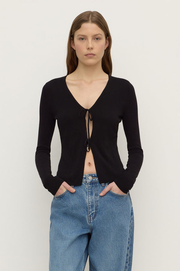 A woman wearing a black long sleeve knit top with two self tie closure at the front, with blue denim jeans, standing against a plain white background.