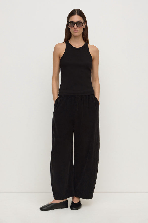 A woman wearing a black sleeveless tank top and barrel-leg black cord pants, paired with black ballet flats and sunglasses, against a plain white background.
