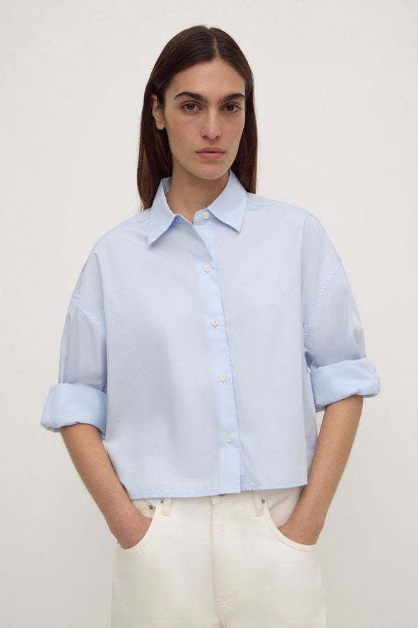 A woman wearing a light blue cropped button-up shirt with rolled sleeves and white jeans against a plain white background.