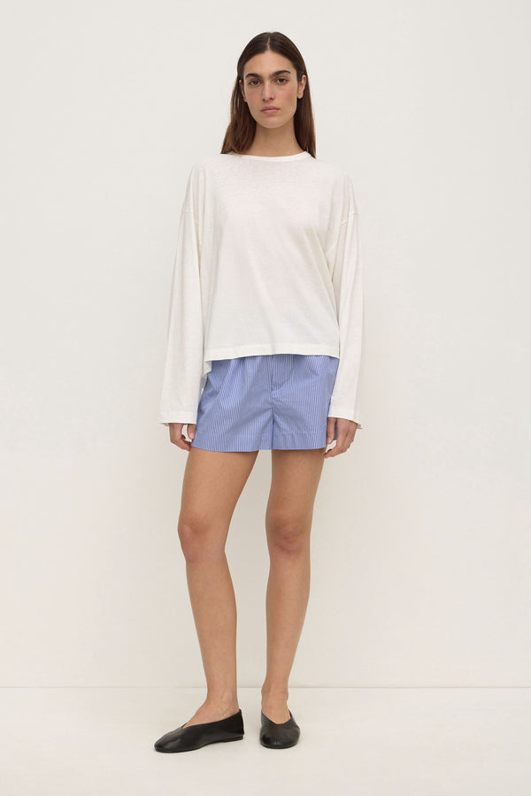 A woman wearing a white long-sleeve top,light blue stripe shorts and black ballet flats against a plain white background.