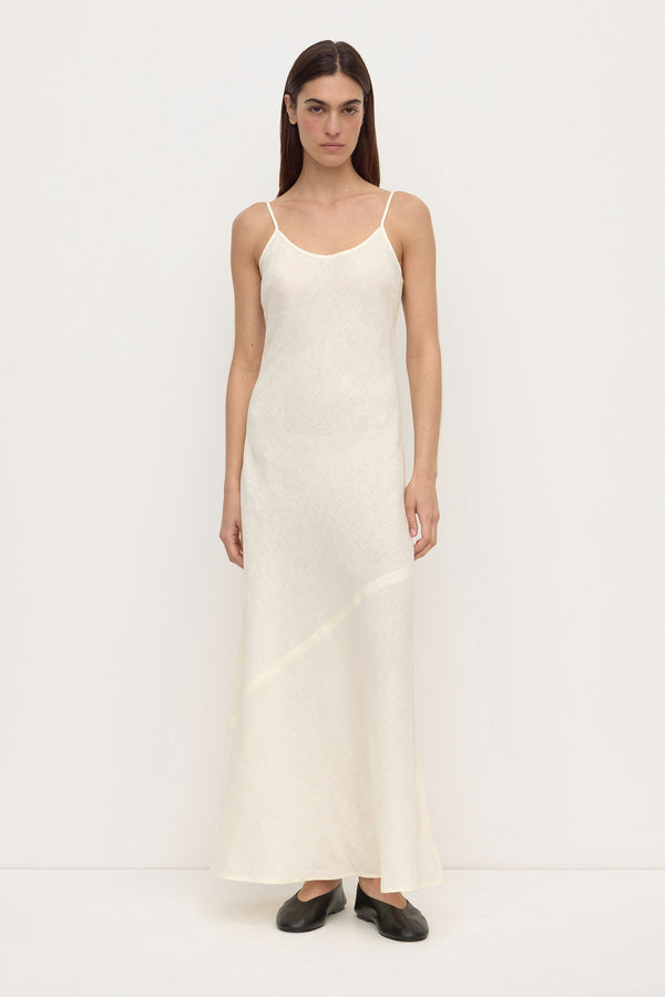 A woman wearing a long, sleeveless cream linen slip dress with thin spaghetti straps and a diagonal seam detail near the lower skirt, paired with black ballet flats, set against a plain white background.