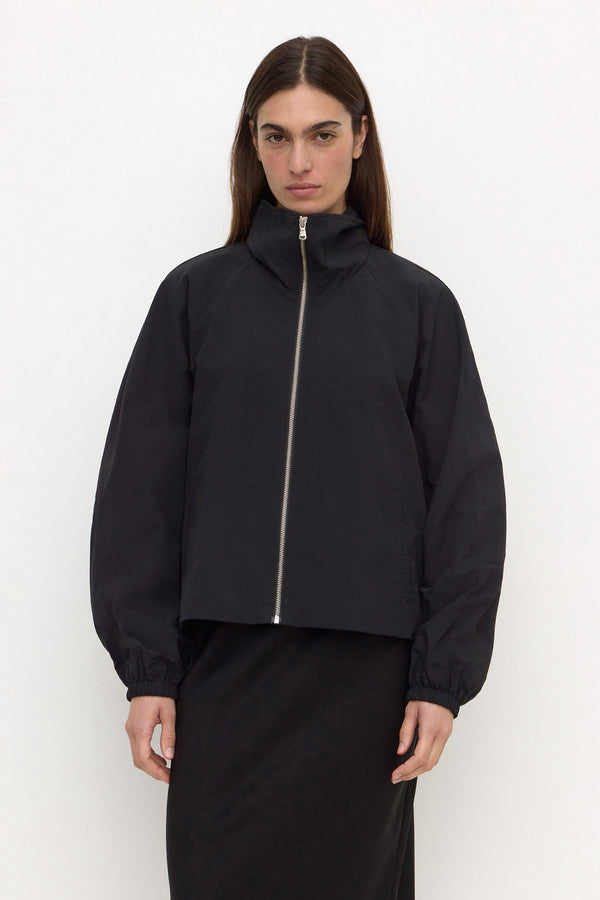 A woman wearing a black cropped zip-up nylon jacket with a high stand collar and elasticised cuffs, paired with a black skirt, against a plain white background.