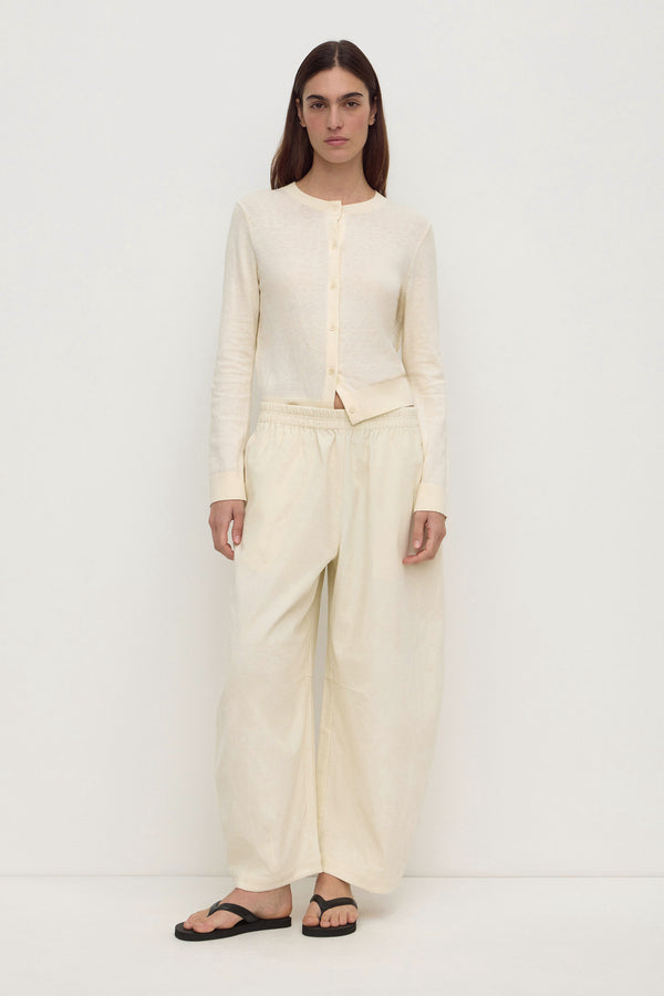 A woman wearing a cream long-sleeve button-up cropped cardigan and cream barrel-leg pants, paired with black flip-flop sandals against a plain white background.