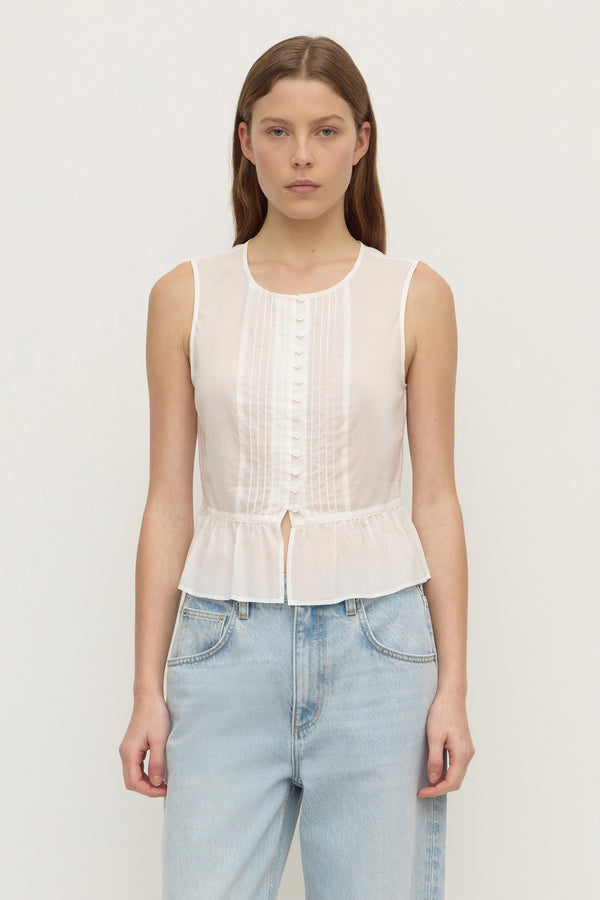 A woman wearing a sleeveless white blouse with vertical button details and a ruffled hem, paired with light blue jeans, against a plain off-white background.